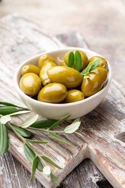 A white ceramic bowl filled with fresh green olives decorated with olive leaves, selected for high-quality olive oil export.