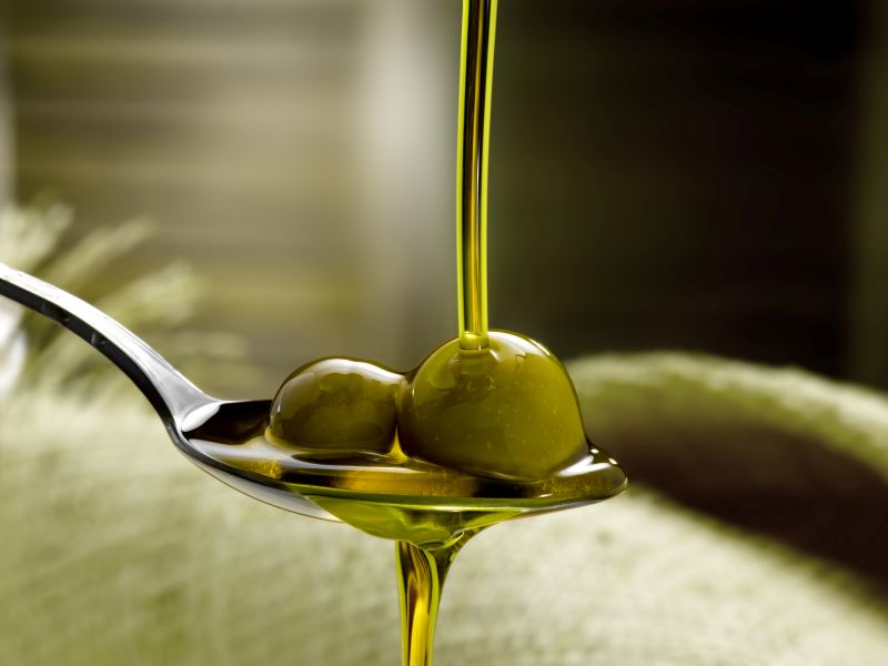 Um fio contínuo de azeite de oliva extra virgem dourado sendo despejado sobre azeitonas verdes em uma colher de metal.
