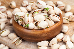 A wooden bowl filled with roasted, split pistachios on a wooden surface.