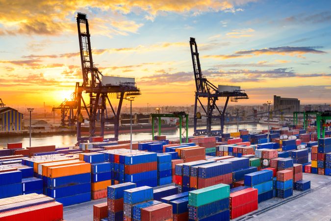 Large harbor cranes and colorful export and shipping containers at a busy commercial port during sunset.